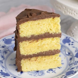 A piece of three layer yellow cake with chocolate frosting on a blue and white plate.