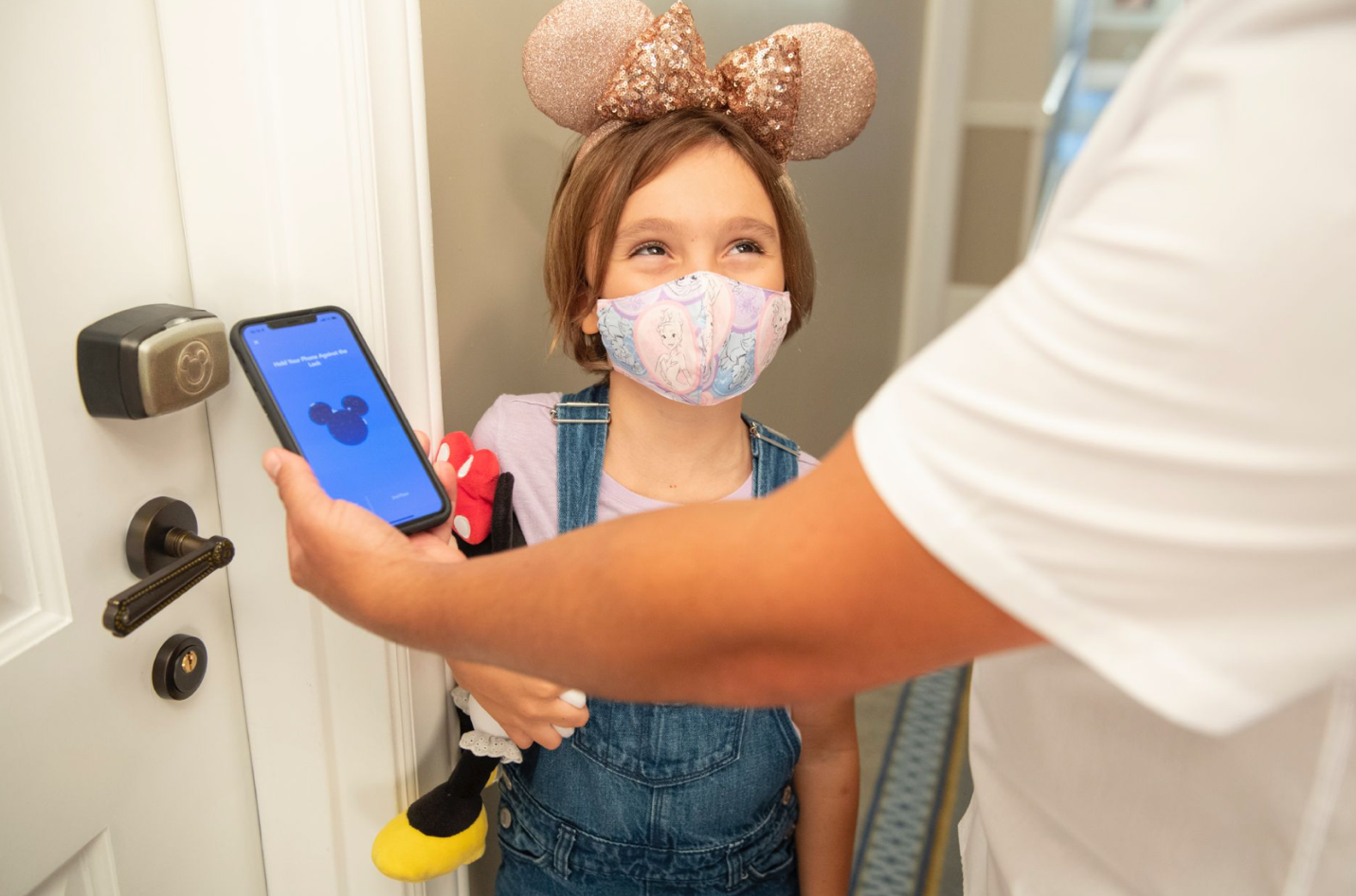 A little girl wearing Minnie Mouse Ears smiles as dad uses his mobile phone to unlock their Disney Resort hotel room door.