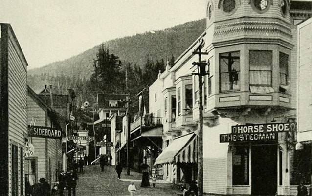 Black and white photo of a street lined with  shops with mountains in background. 